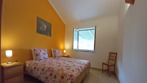 Une chambre à coucher comprenant un lit, deux tables de chevet, une chaise, une fenêtre et un tableau accroché au mur.