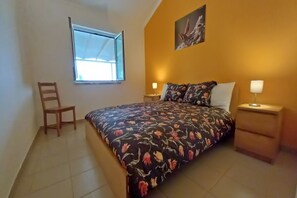 Une chambre à coucher avec un lit, une chaise, une table de chevet, une lampe, une fenêtre et un tableau au mur.