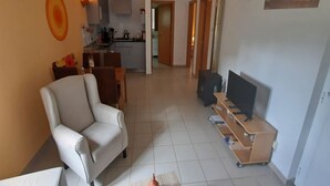 Un salon avec un fauteuil blanc, un téléviseur posé sur un meuble en bois, et une cuisine en arrière-plan.