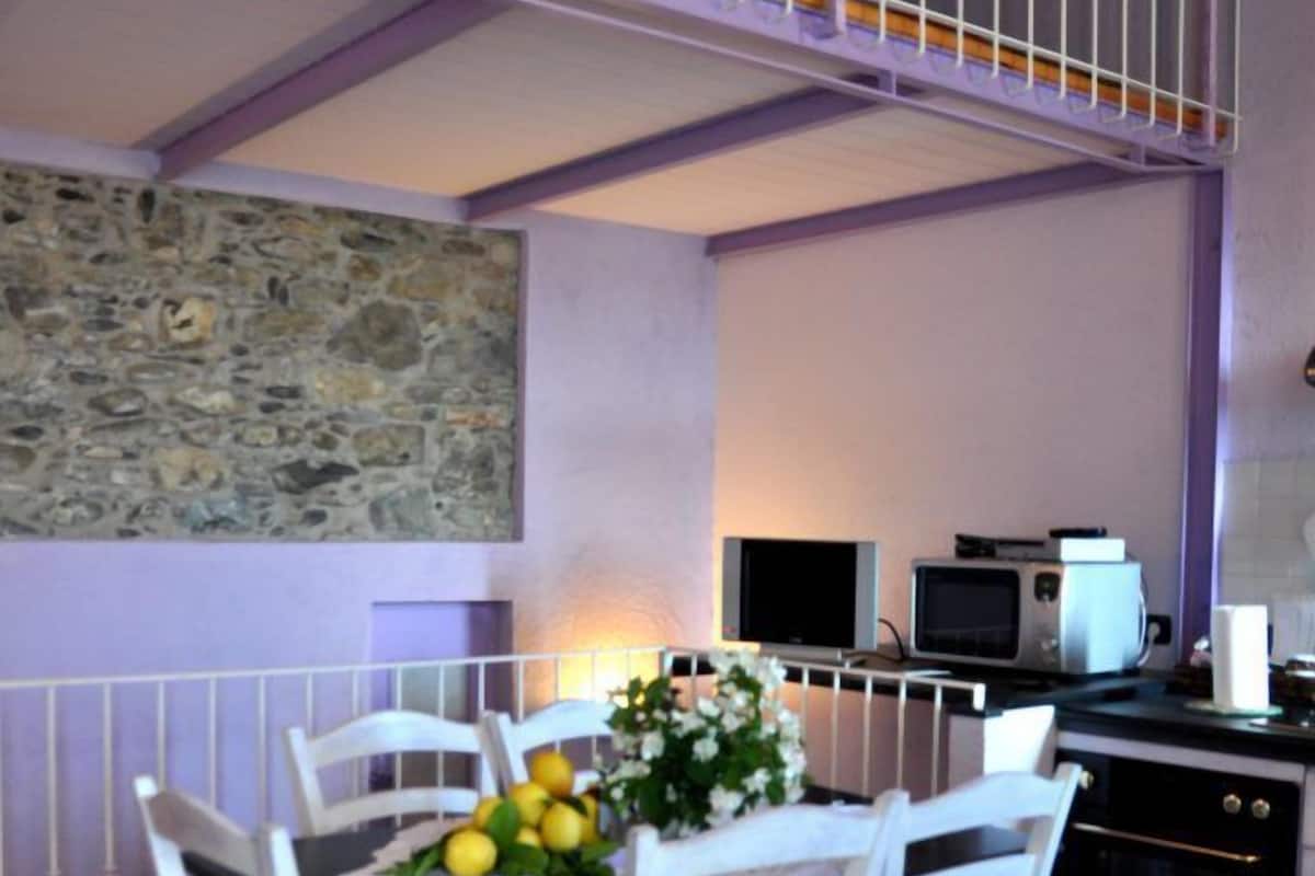 A dining area with a table set for four, surrounded by white chairs, a stone wall, and a kitchen area with appliances.