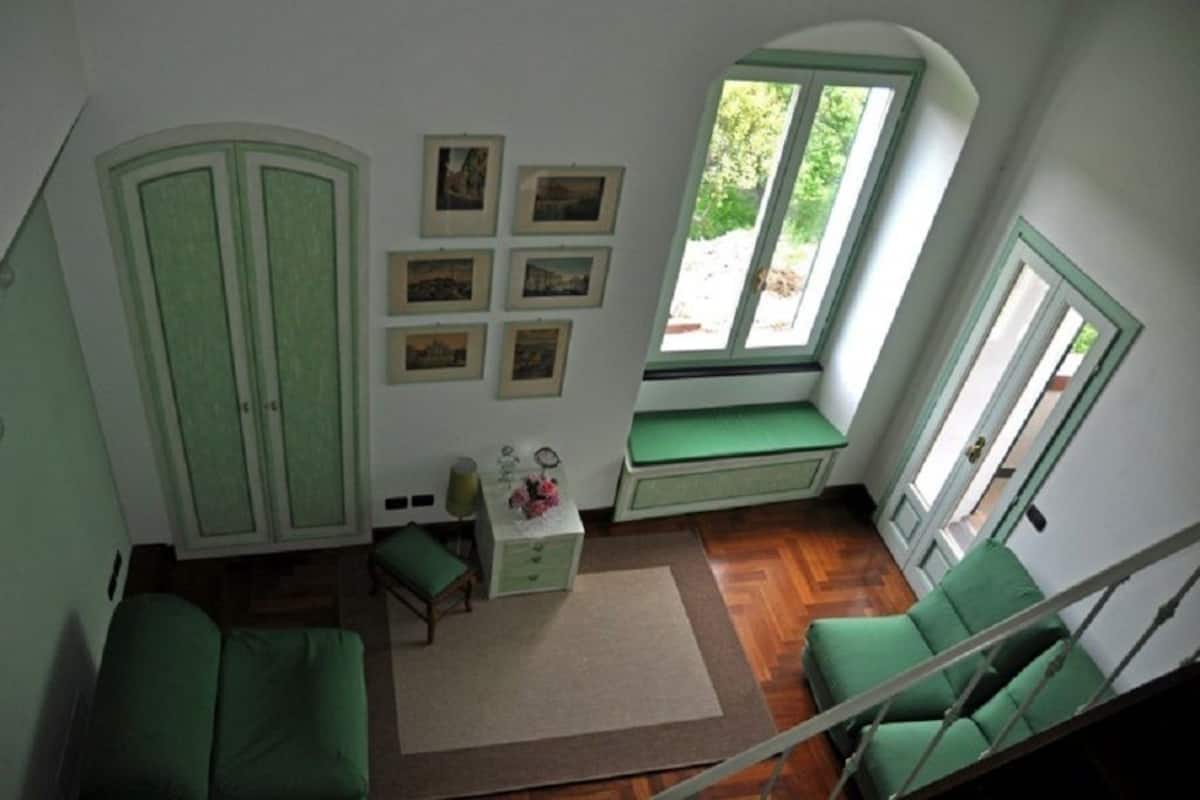 A room with a green sofa, a small table with a lamp, a window seat, and a view of greenery outside.