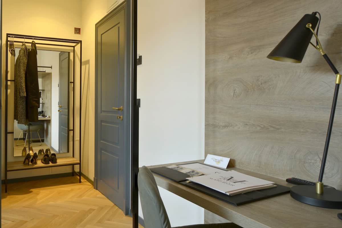 A hotel room with a wooden floor, a desk with a lamp, a mirror, and a coat rack with shoes.