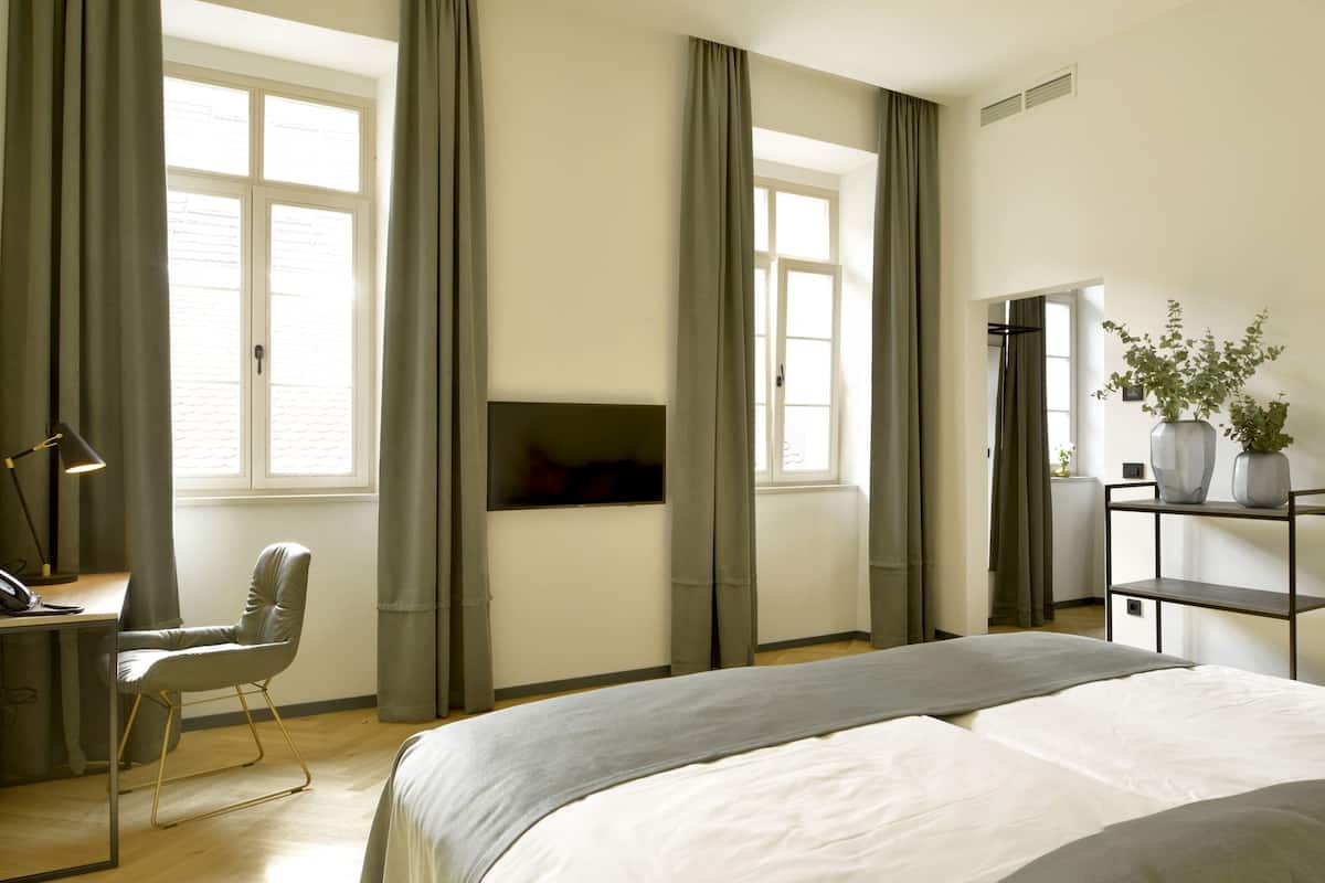 A modern hotel room with a large bed, a desk with a chair, a TV mounted on the wall, and a shelf with plants.