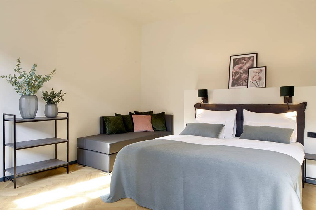 A bedroom with a bed, a bench, a shelf with plants, and two wall-mounted lamps.