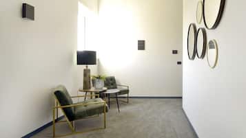 A modern hotel room with a grey carpet, two green upholstered chairs, a small round table, a floor lamp, and a wall with multiple mirrors.