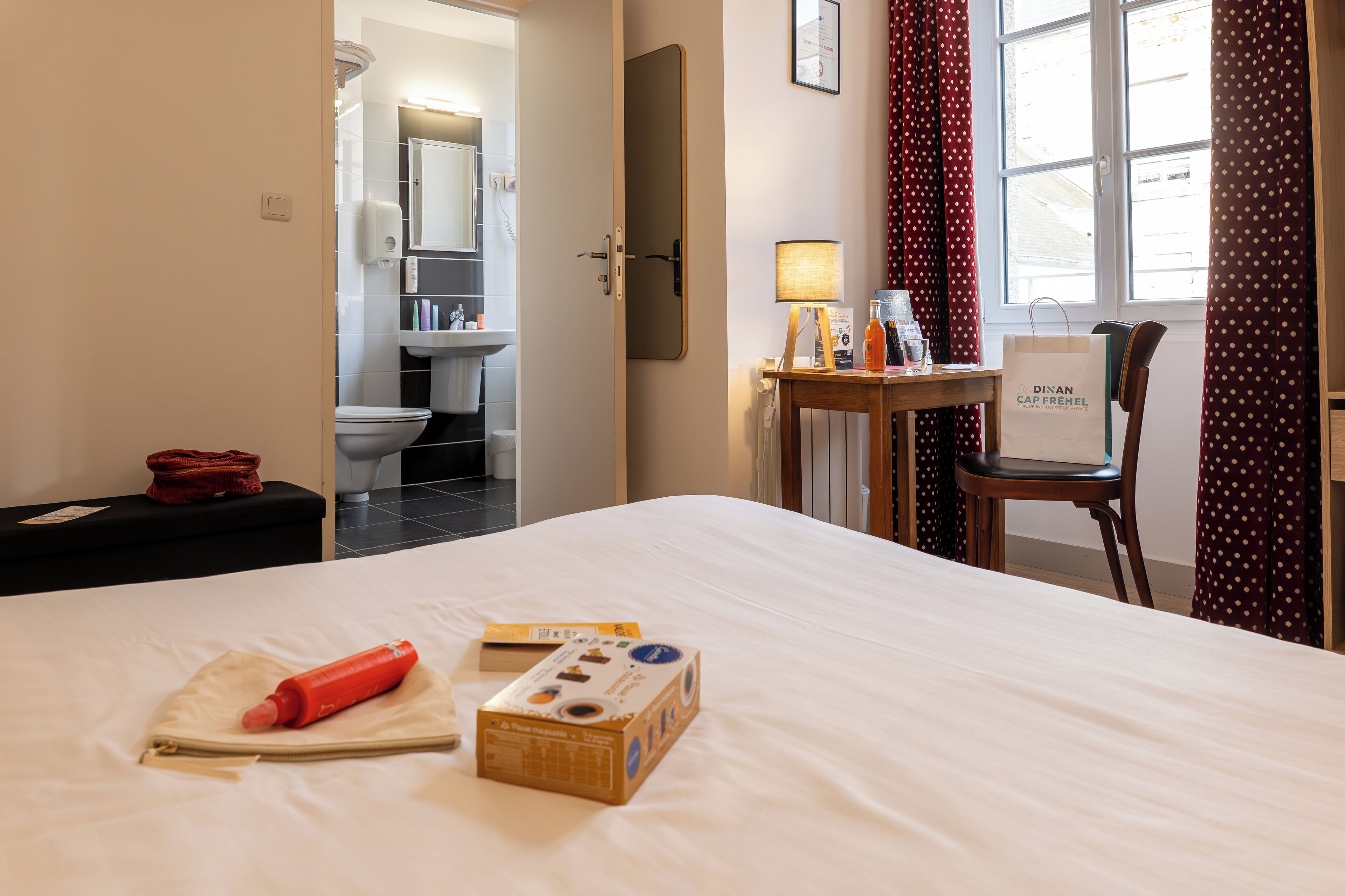 A hotel room with a bed, a bedside table with a lamp, a chair, a suitcase, and a bathroom visible through an open door.