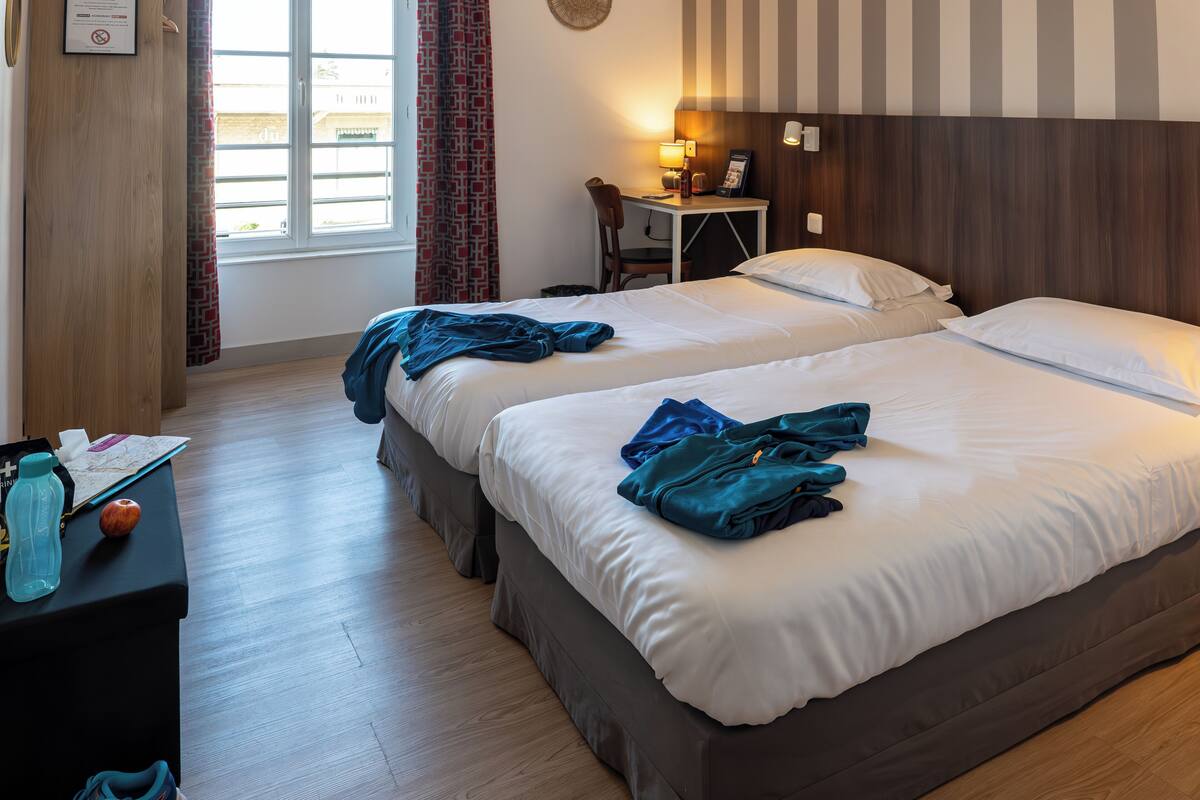 Ein Hotelzimmer mit zwei Betten, einem Schreibtisch mit Stuhl, einer Lampe und Blick ins Freie durch ein Fenster.