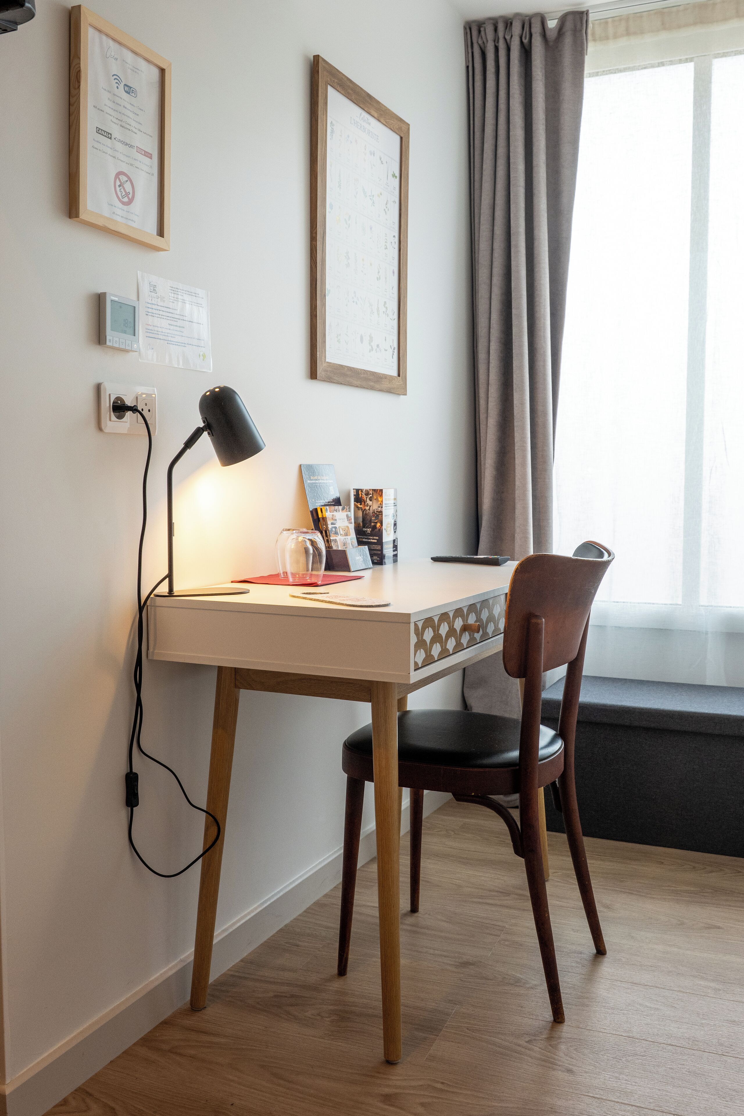 Een klein, netjes ingerichte werkplek met een bureau, stoel en een lamp. Er hangen ingelijste diploma’s aan de muur, een raam met gordijnen en een stopcontact.