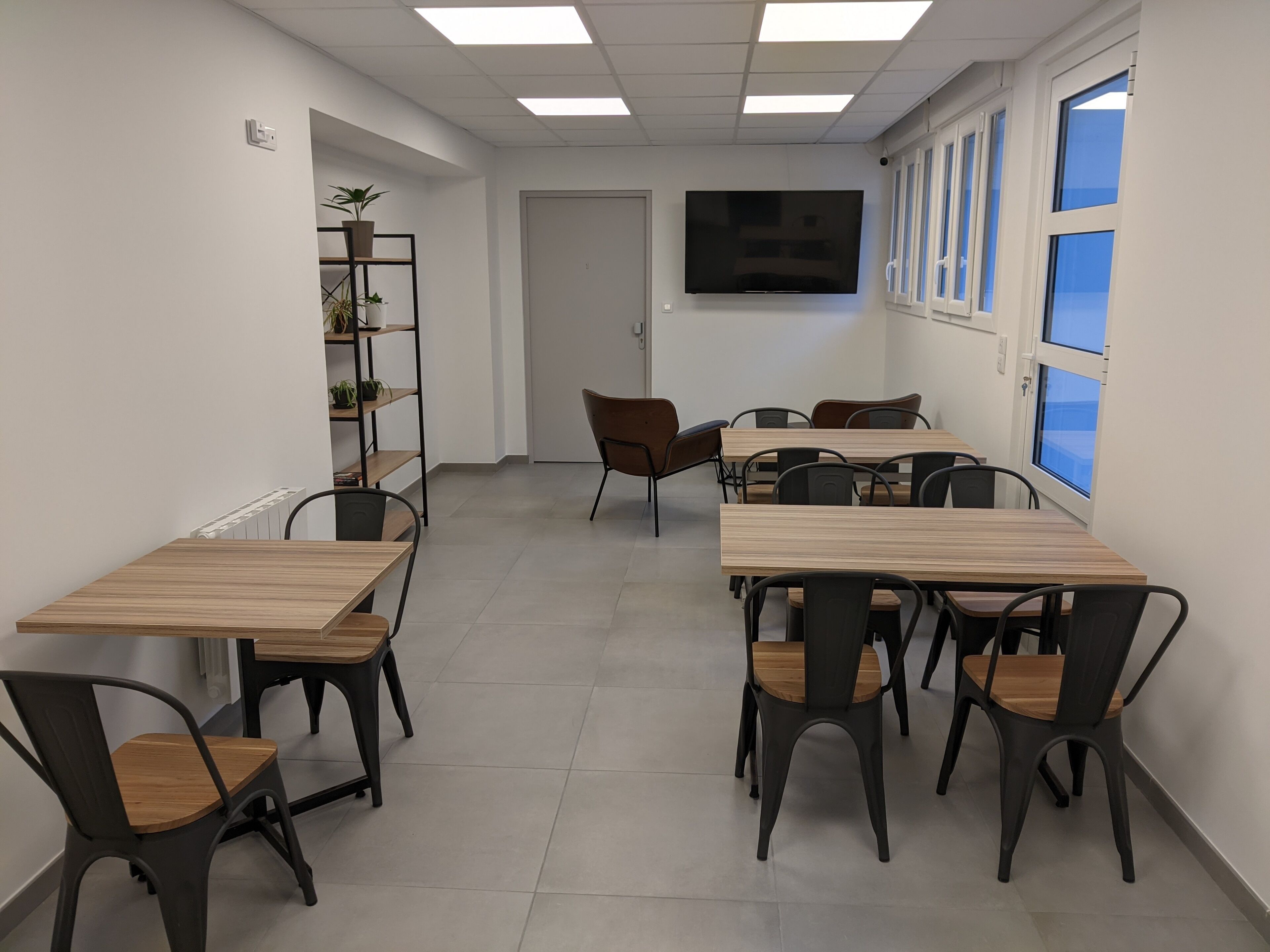 A conference room with multiple tables and chairs, a large flat-screen TV on the wall, and a window with blinds.