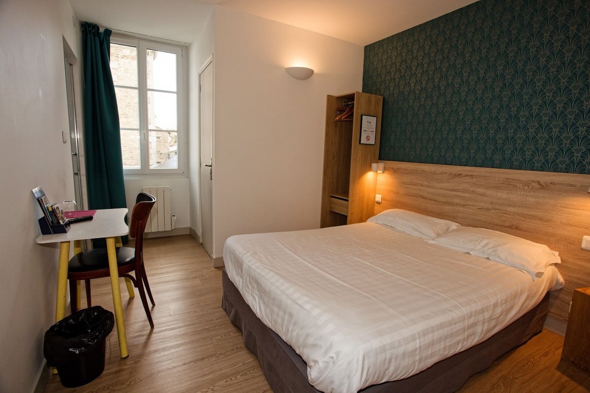Ein Hotelzimmer mit einem Bett, einem Schreibtisch mit Stuhl, einem Heizkörper und einem Fenster mit Vorhängen.