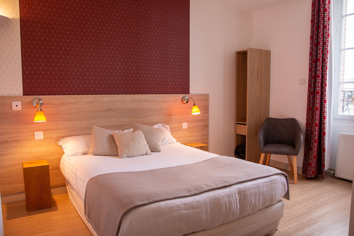 Ein ordentlich bezogenes Bett mit Kopfteil, zwei Nachttischlampen, ein Sessel und ein Kleiderschrank in einem Zimmer mit gemustertem Wandpapier und einem Vorhangfenster.
