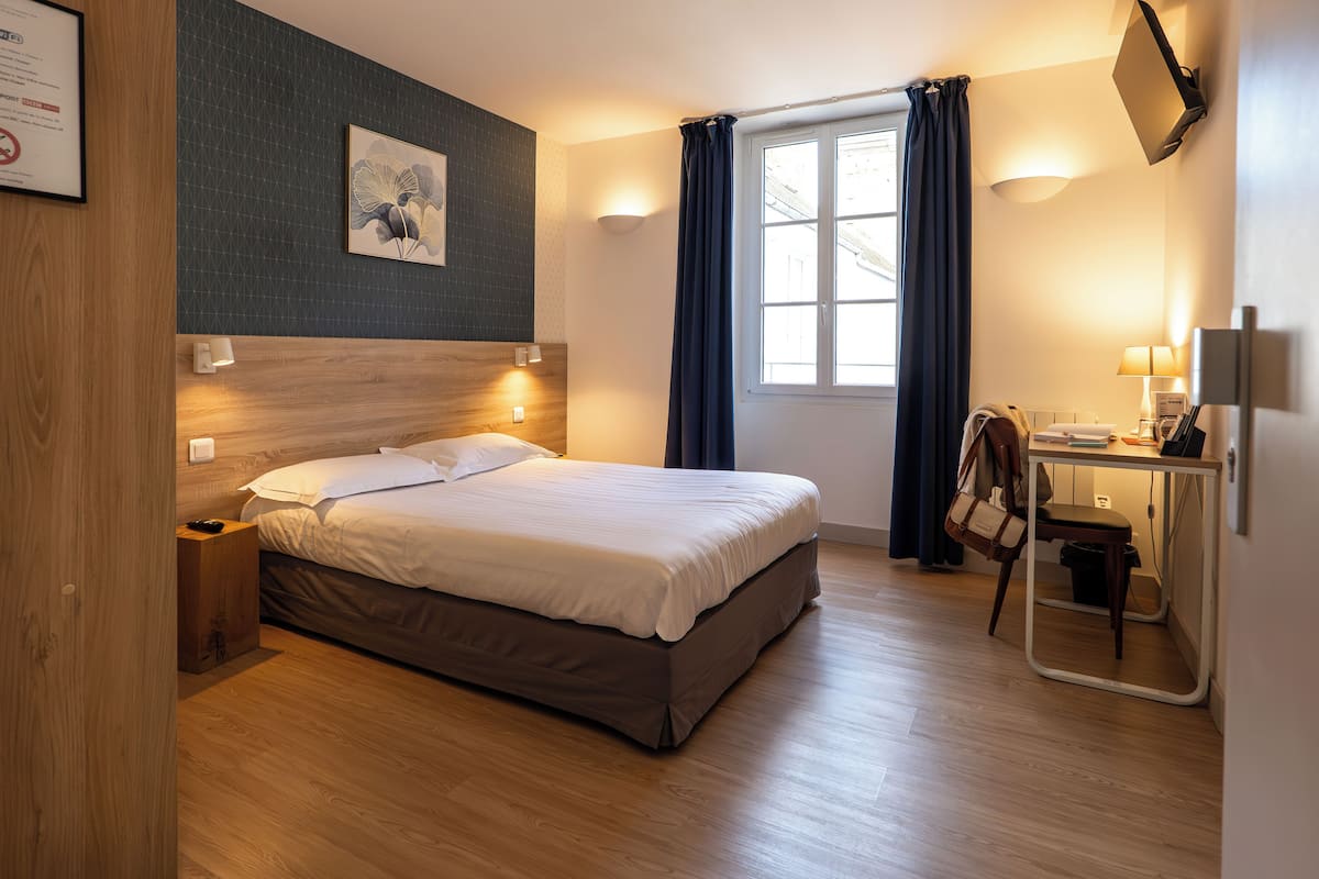 Ein Hotelzimmer mit einem Bett, Nachttischlampen, einem Schreibtisch mit Stuhl, einem Fenster mit Vorhängen und einem an der Wand befestigten Fernseher.