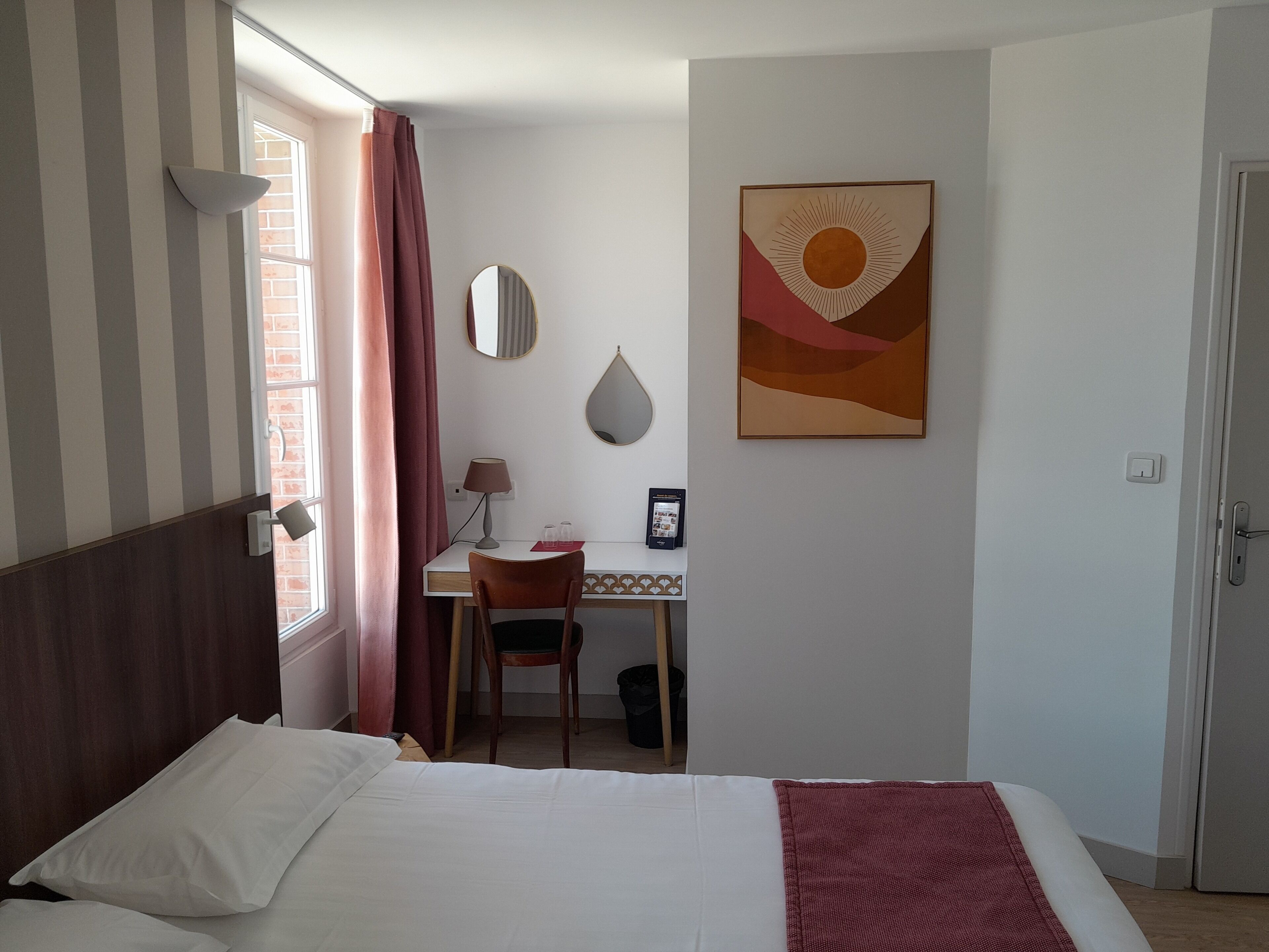 Hotellihuone, jossa on sänky, työpöytä, tuoli, ikkuna verhoilla, seinälle kiinnitetty peili ja kehystetty taulu.