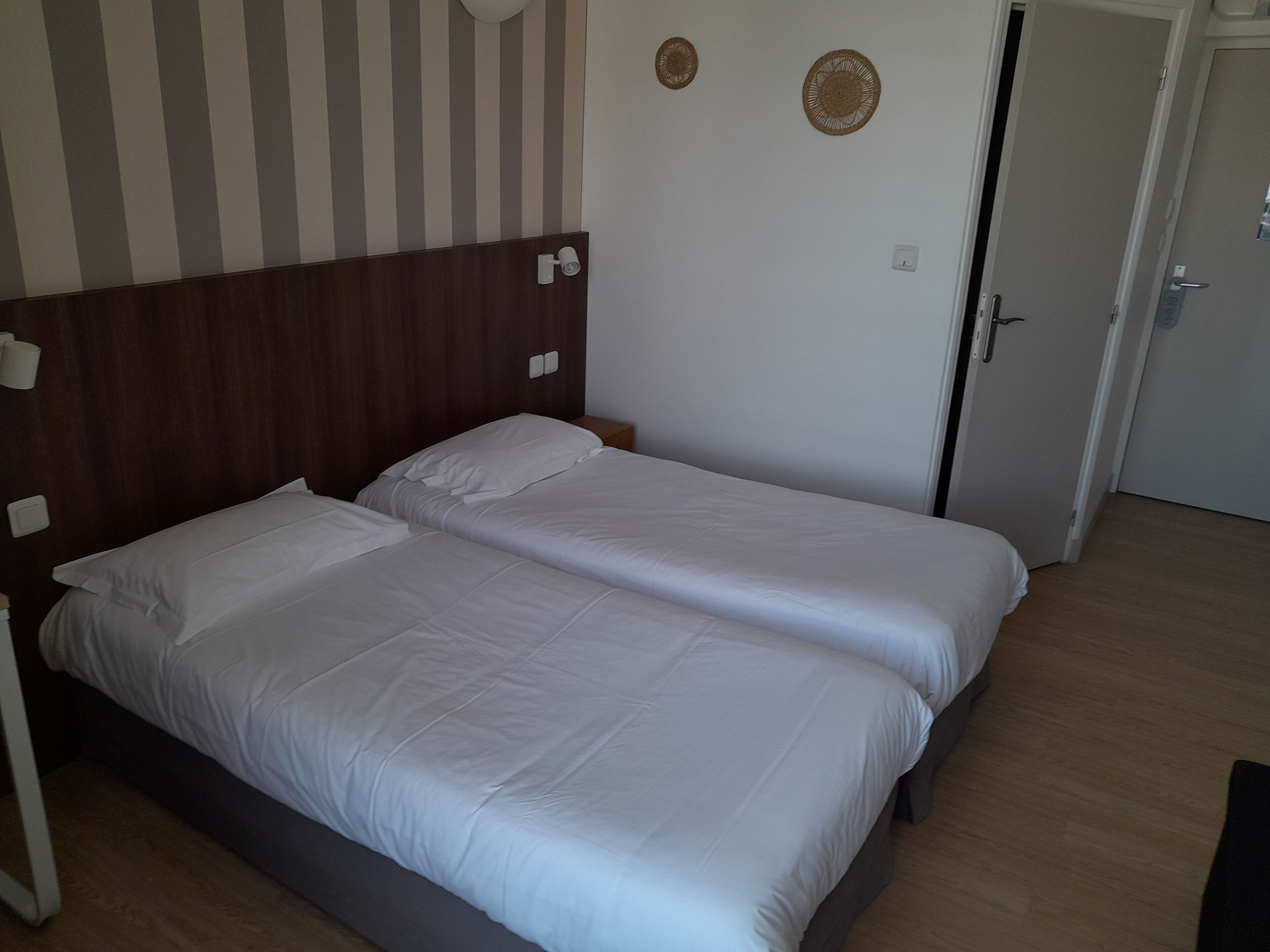 Un lit double avec une literie blanche, dans une chambre au parquet en bois et dont un mur est rayé.