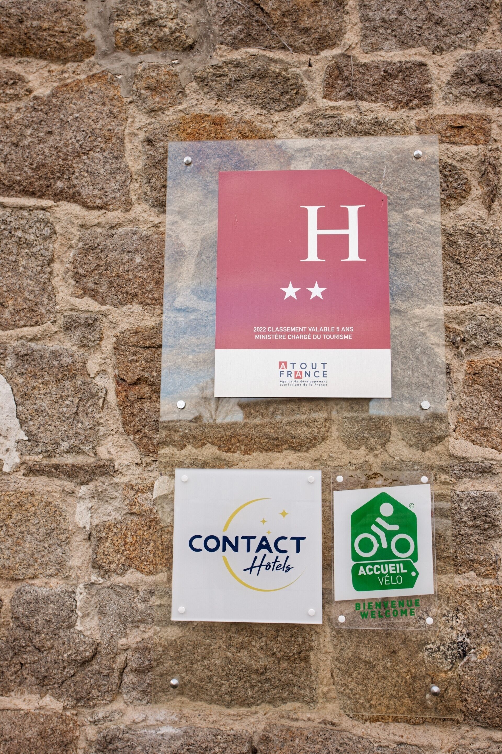 A wall with multiple signs, including a red sign with a white 'H' and stars, a white sign with 'CONTACT Hotels', and a green sign with 'ACCEIL VÉLO'.