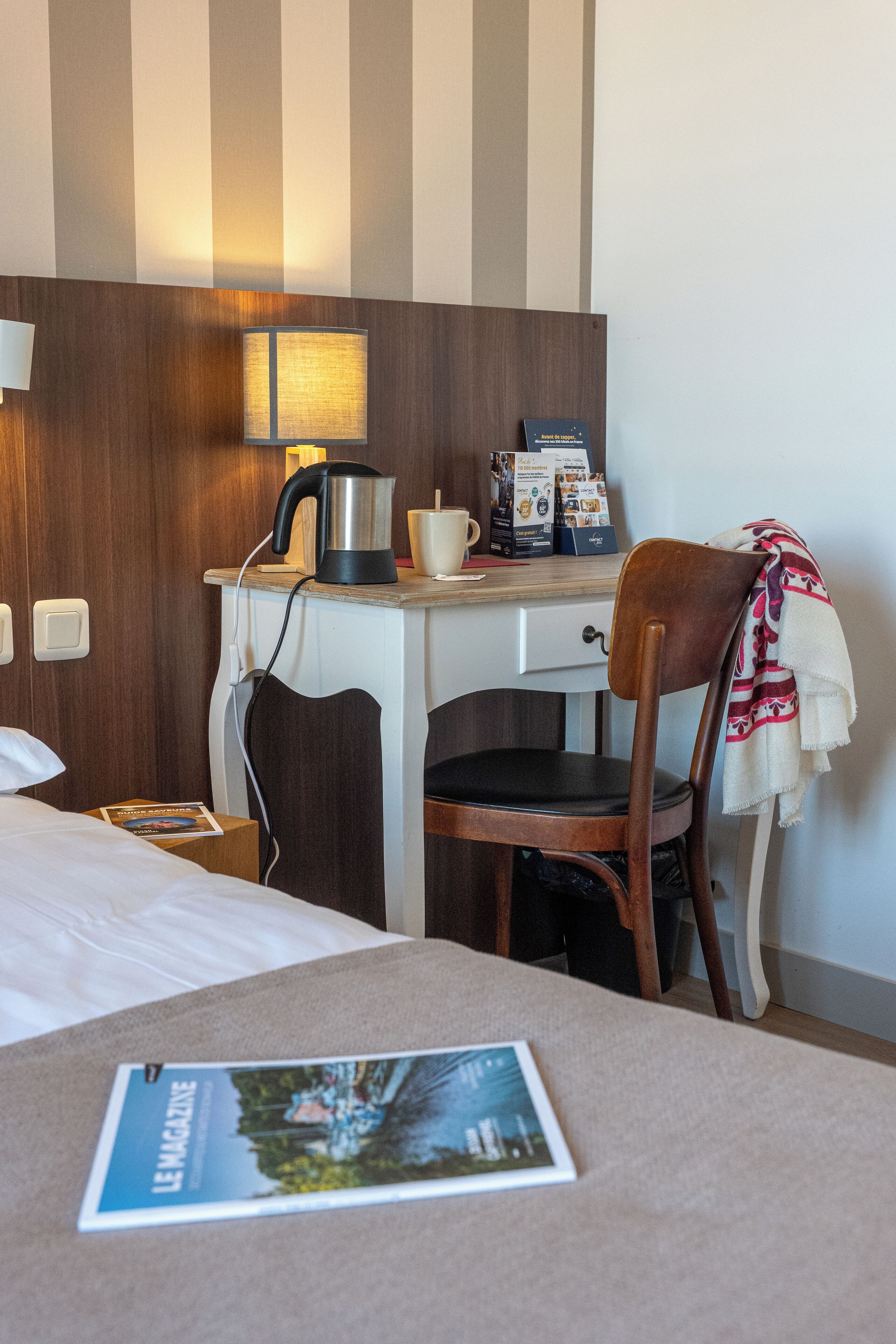 Habitació d'hotel amb un llit, una taula de nit amb una cafetera, una cadira i una revista damunt del llit.