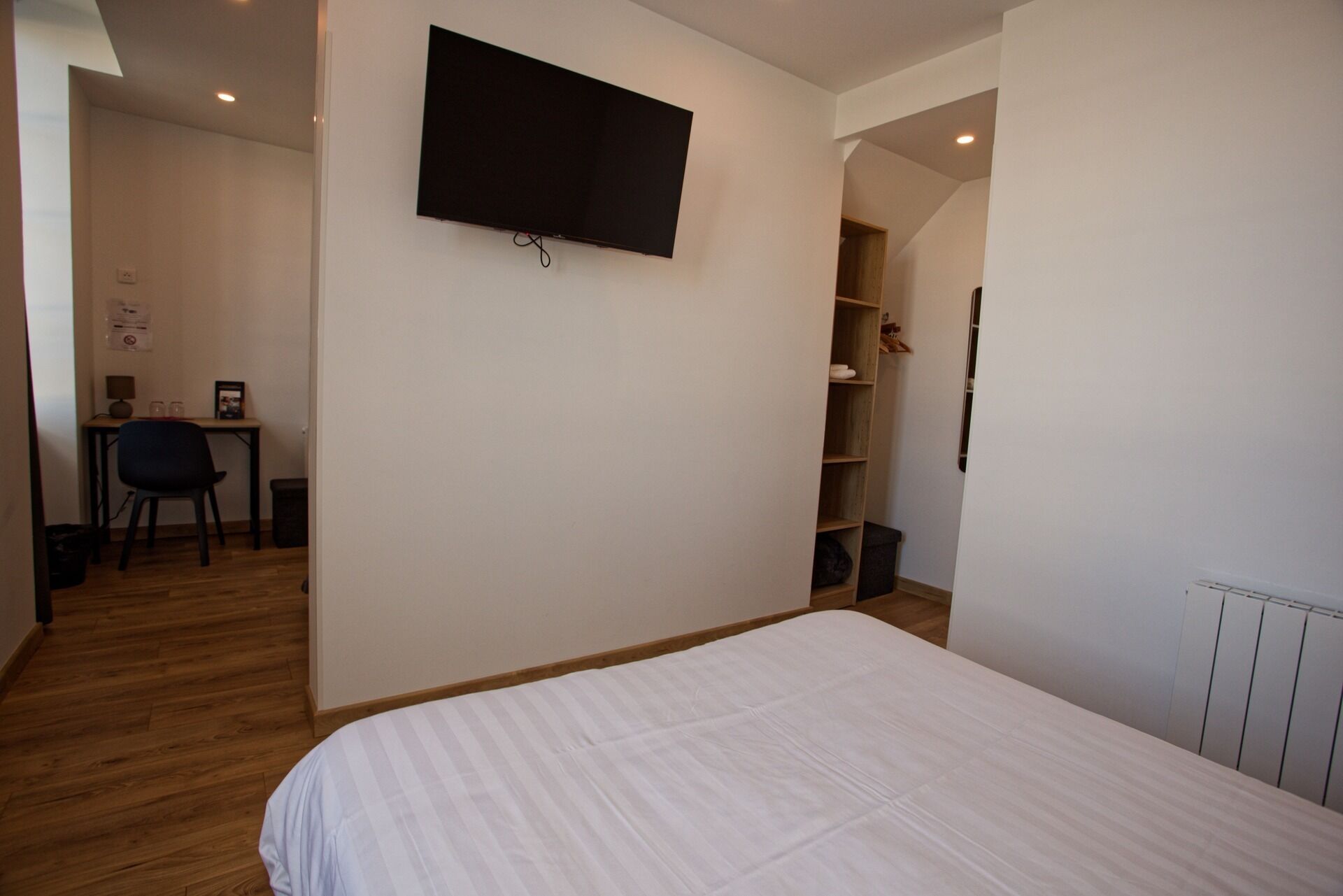 Una camera da letto con un letto, una scrivania con sedia, una televisione a parete e una scaffalatura.