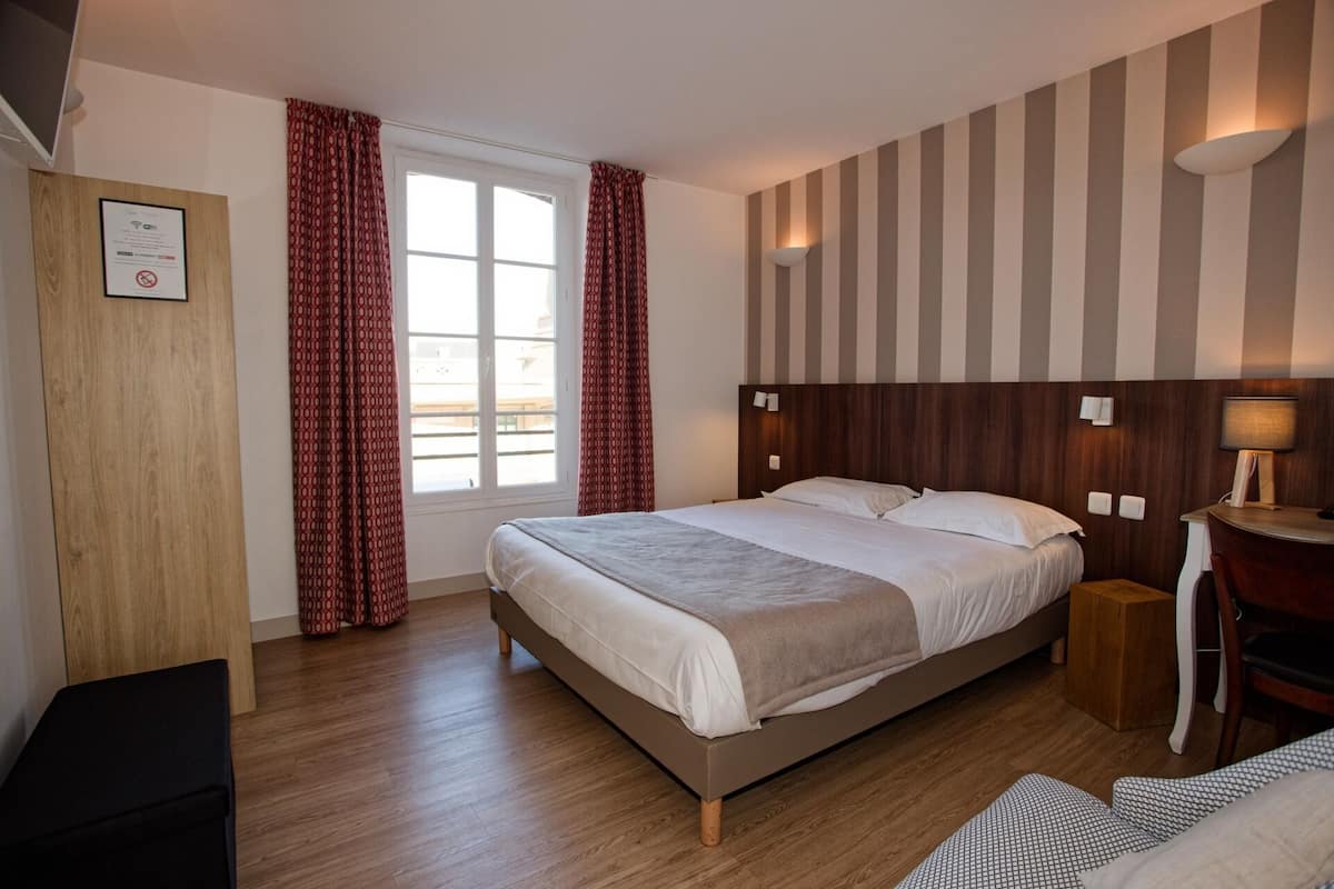 Ein Hotelzimmer mit einem Bett, einem Schreibtisch und einem Stuhl. Es gibt ein Fenster mit Vorhängen, eine Wandlampe und eine Holztür.