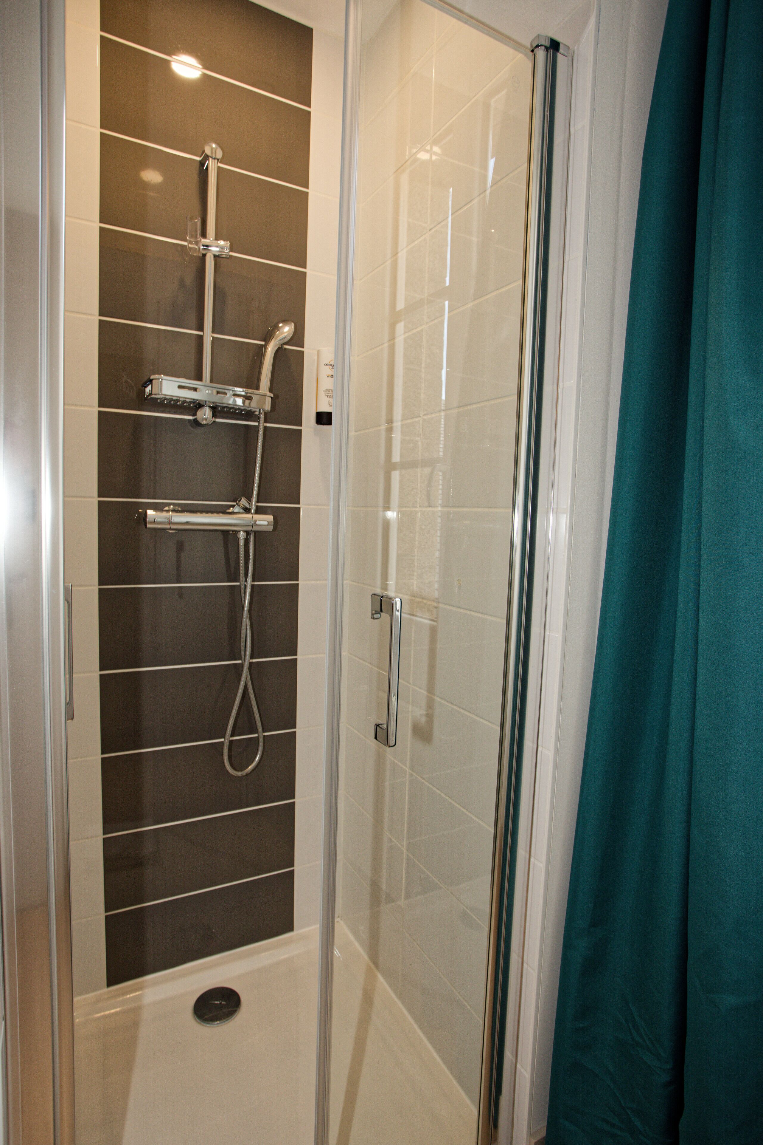 Une cabine de douche fermée par des parois de verre, dotée d’un pommeau de douche et d’un panneau de commande en chrome, de murs recouverts de carreaux blancs et d’un rideau de douche de couleur turquoise.