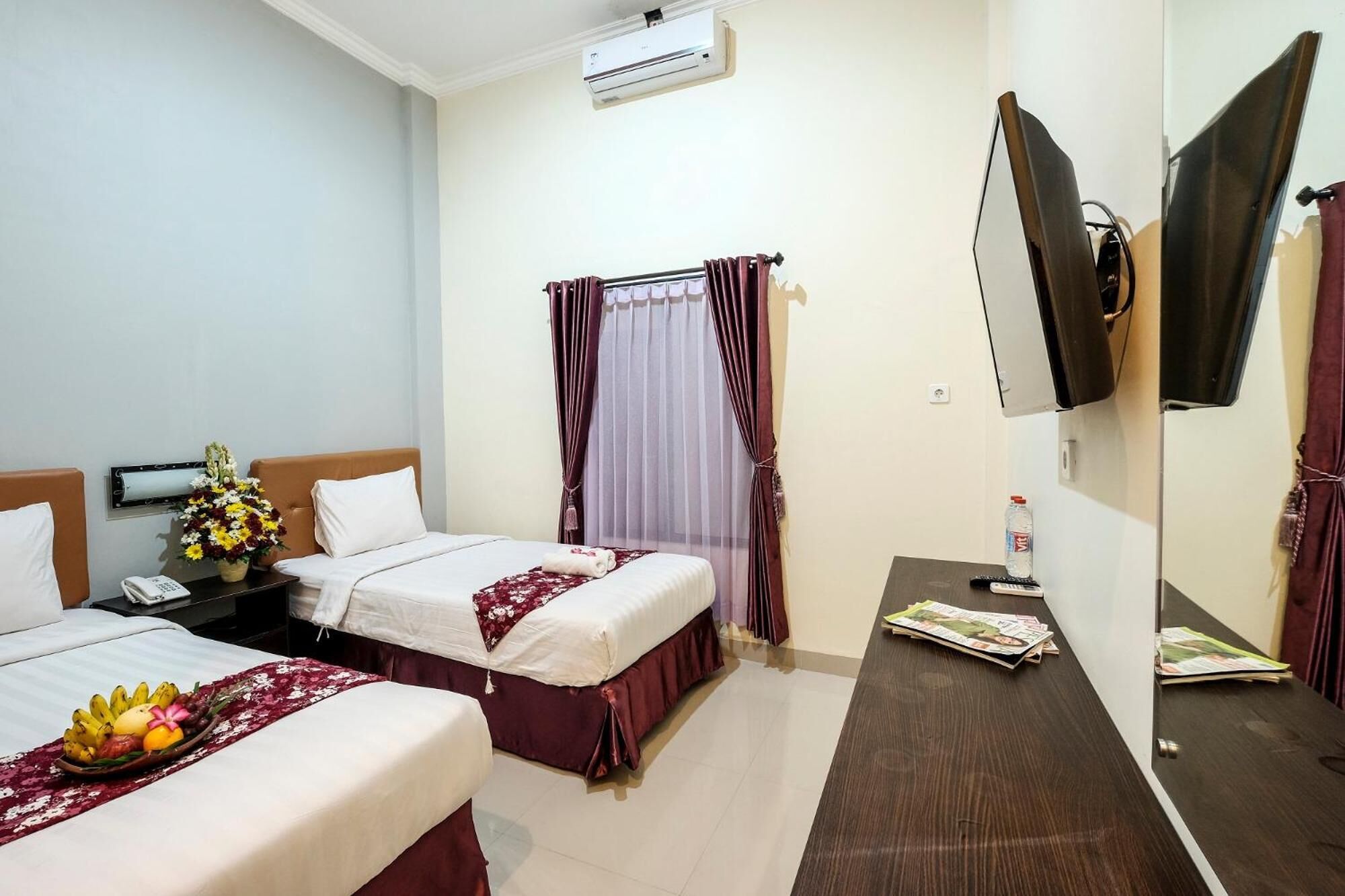 Habitación de hotel con dos camas, una mesita de noche con un teléfono y un jarrón de flores, un escritorio con un espejo y un televisor montado en la pared.