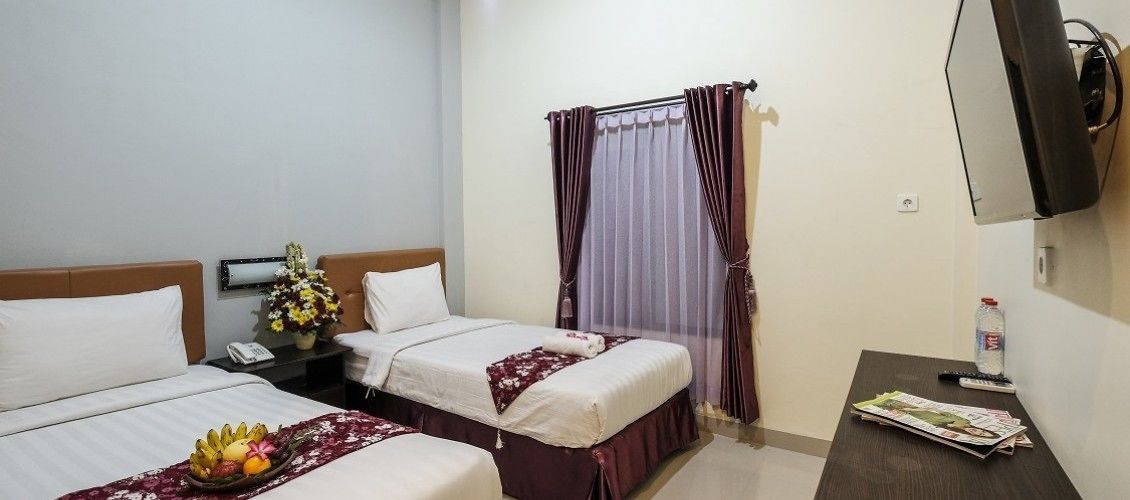 A hotel room with two beds, a bedside table with a phone and a bottle, a TV mounted on the wall, and a window with curtains.