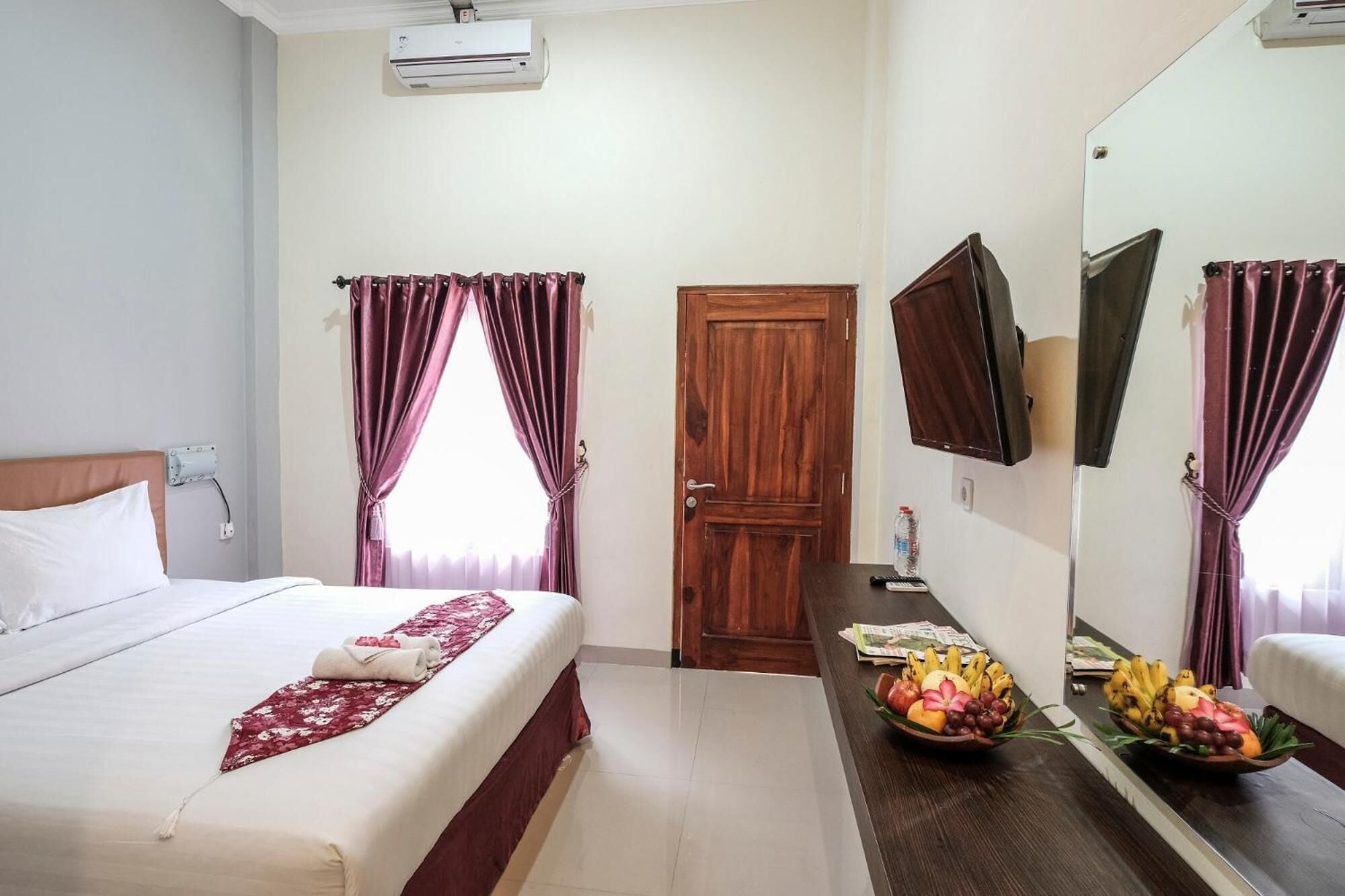 A hotel room with a bed, a wooden door, a mirror, a television, and a fruit bowl on the dresser.