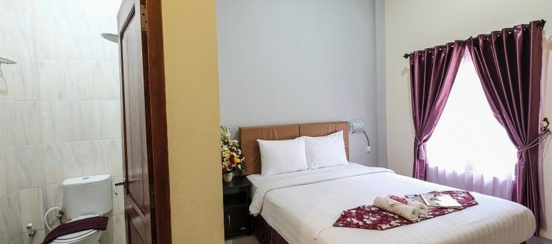 Una camera d'albergo con un letto, un comodino con una lampada, una finestra con tende e un bagno con il water e la doccia.