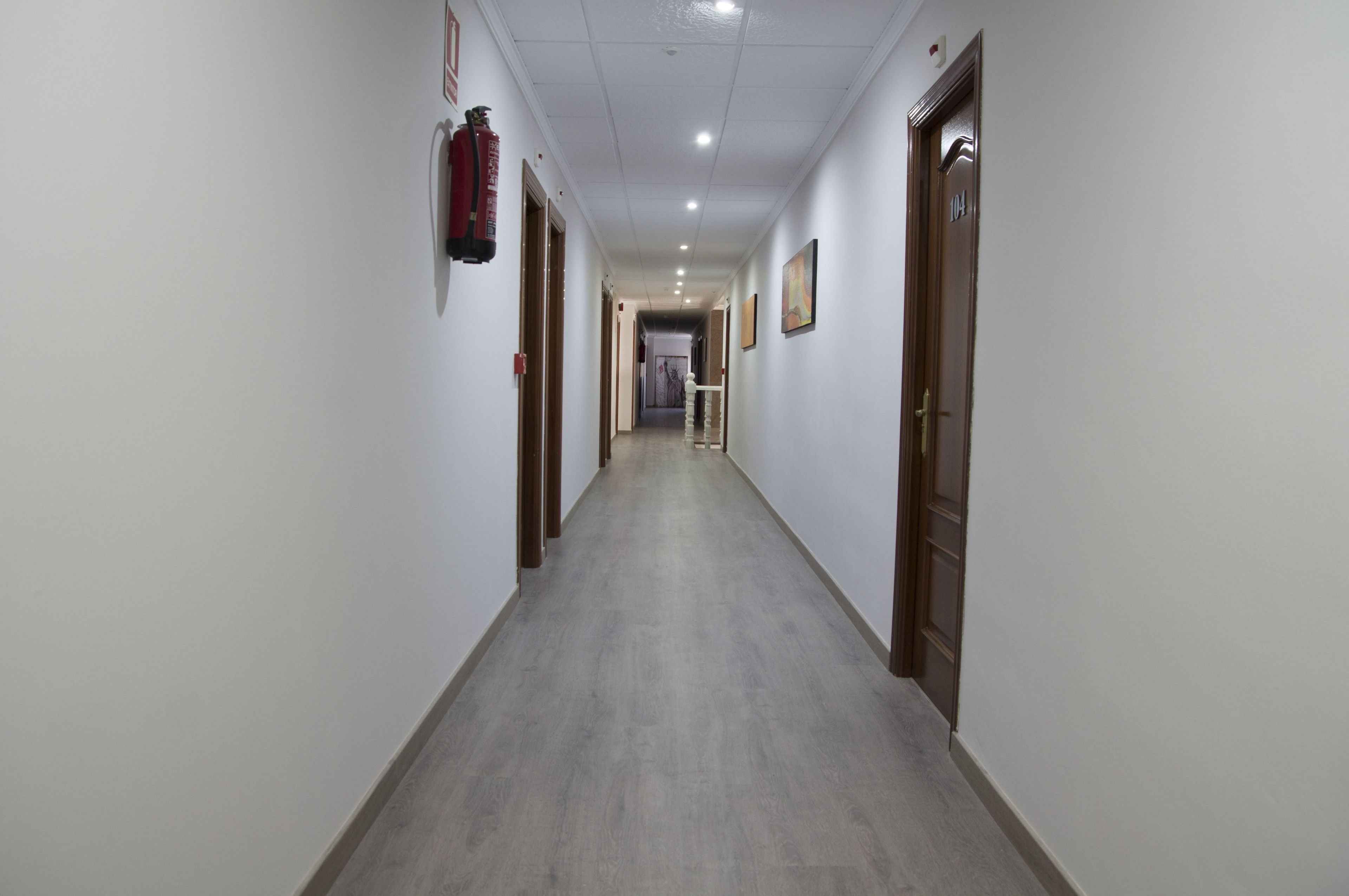 Un largo y estrecho pasillo con paredes blancas y puertas de madera que conduce a una habitación con una cama y un ventanal.