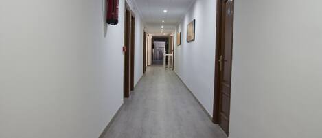 Un largo y estrecho pasillo con paredes blancas y puertas de madera que conduce a una habitación con una cama y un ventanal.