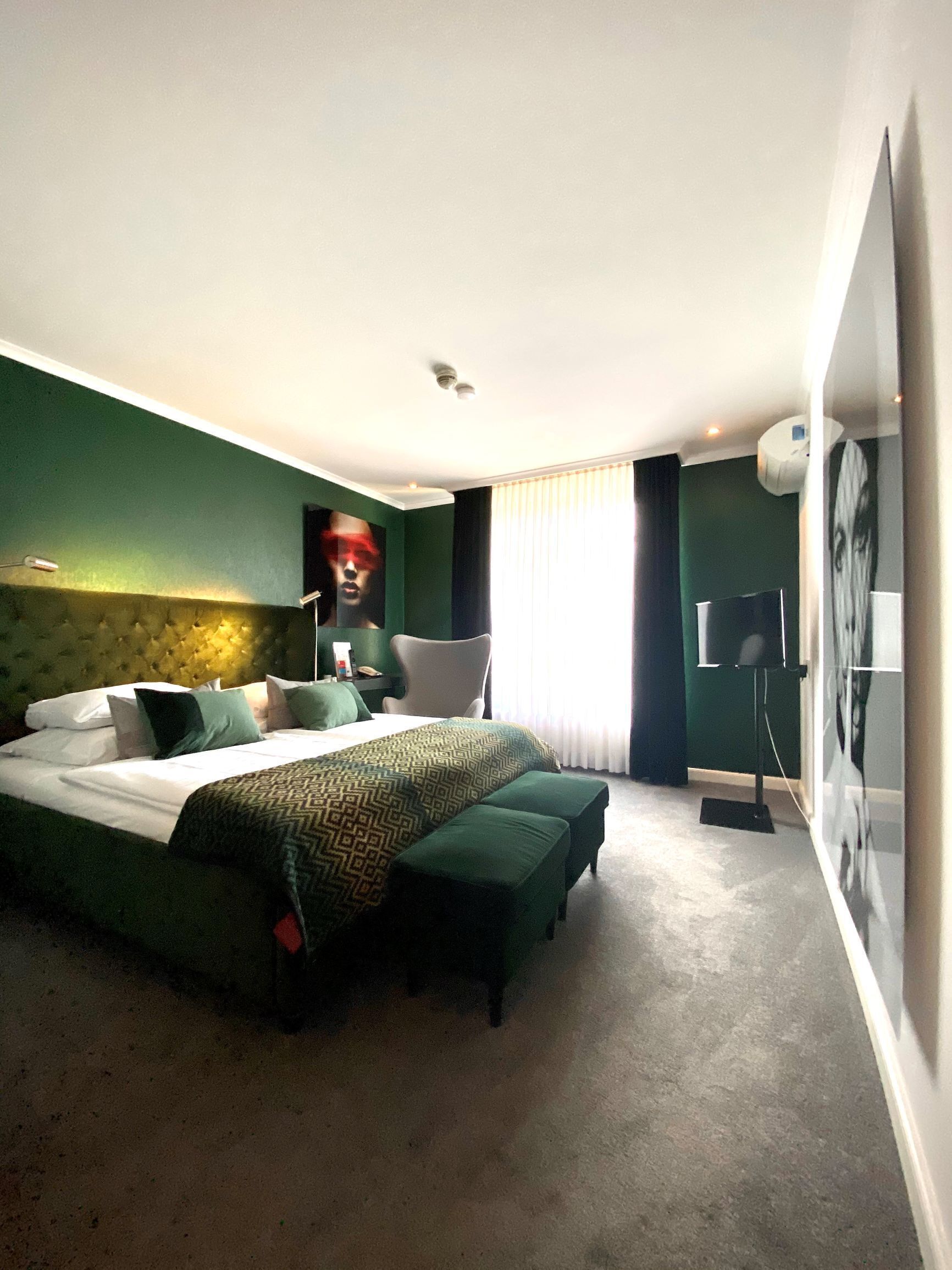 A modern hotel room with a large bed, a tufted headboard, a green accent wall, a large poster, and a standing lamp.