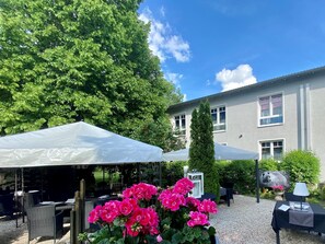 An outdoor seating area with tables and chairs under white canopies, surrounded by greenery and a building with windows. - Parkhotel Bielefeld (Bielefeld)