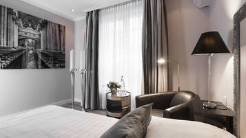 A modern hotel room with a large bed, a seating area with a chair, a side table with a lamp, and a decorative wall piece.