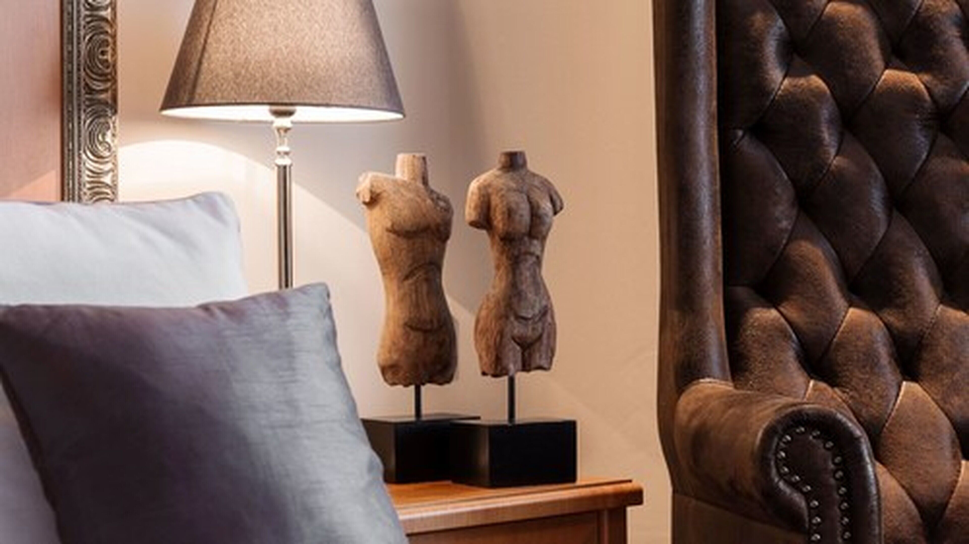 A bedroom with a bed, a lamp, two wooden sculptures, a dark brown leather armchair, and a wooden nightstand.