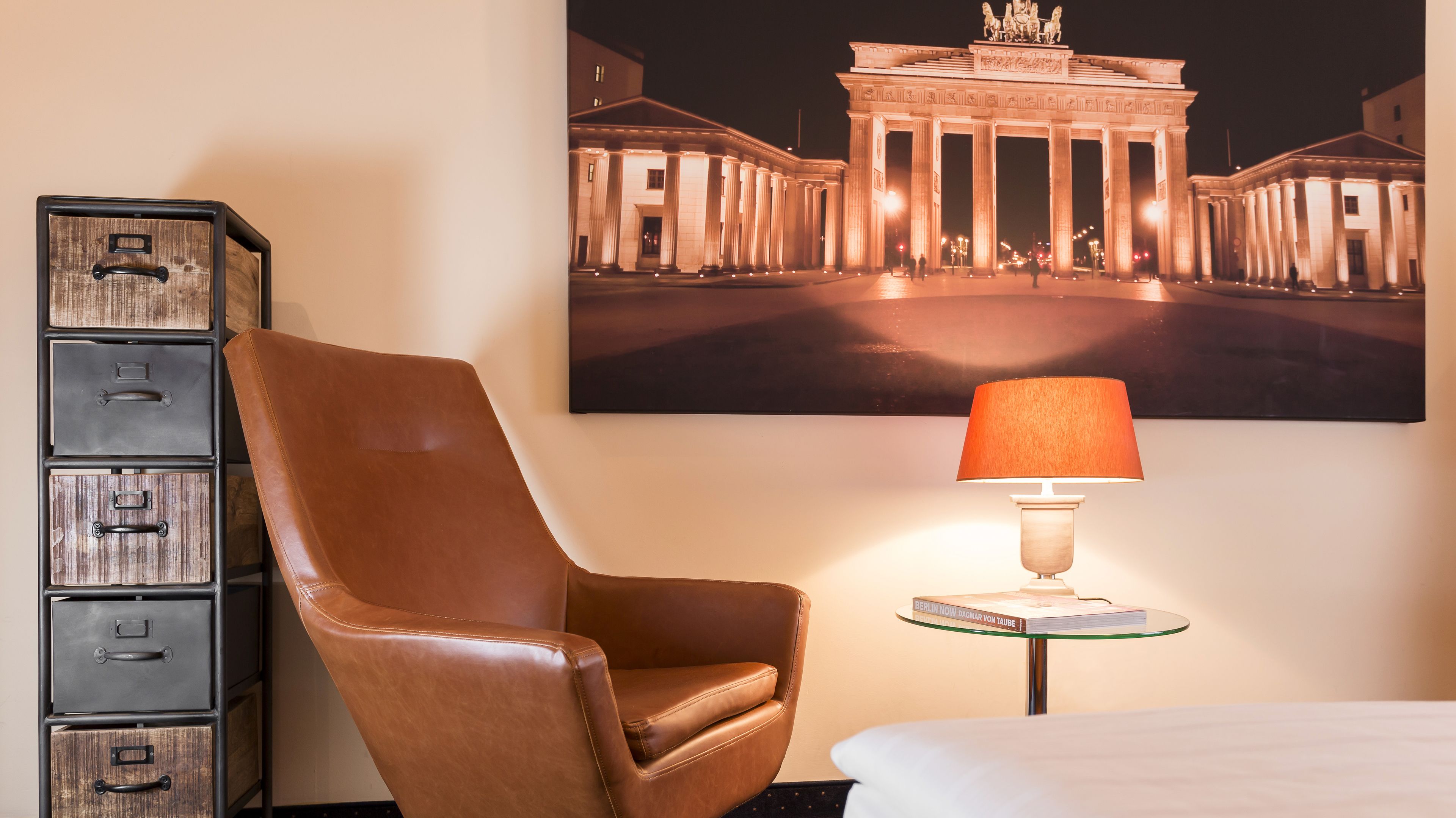 Une chambre d’hôtel avec un fauteuil en cuir, une table en verre avec une lampe, une bibliothèque et un grand tableau encadré représentant un bâtiment.