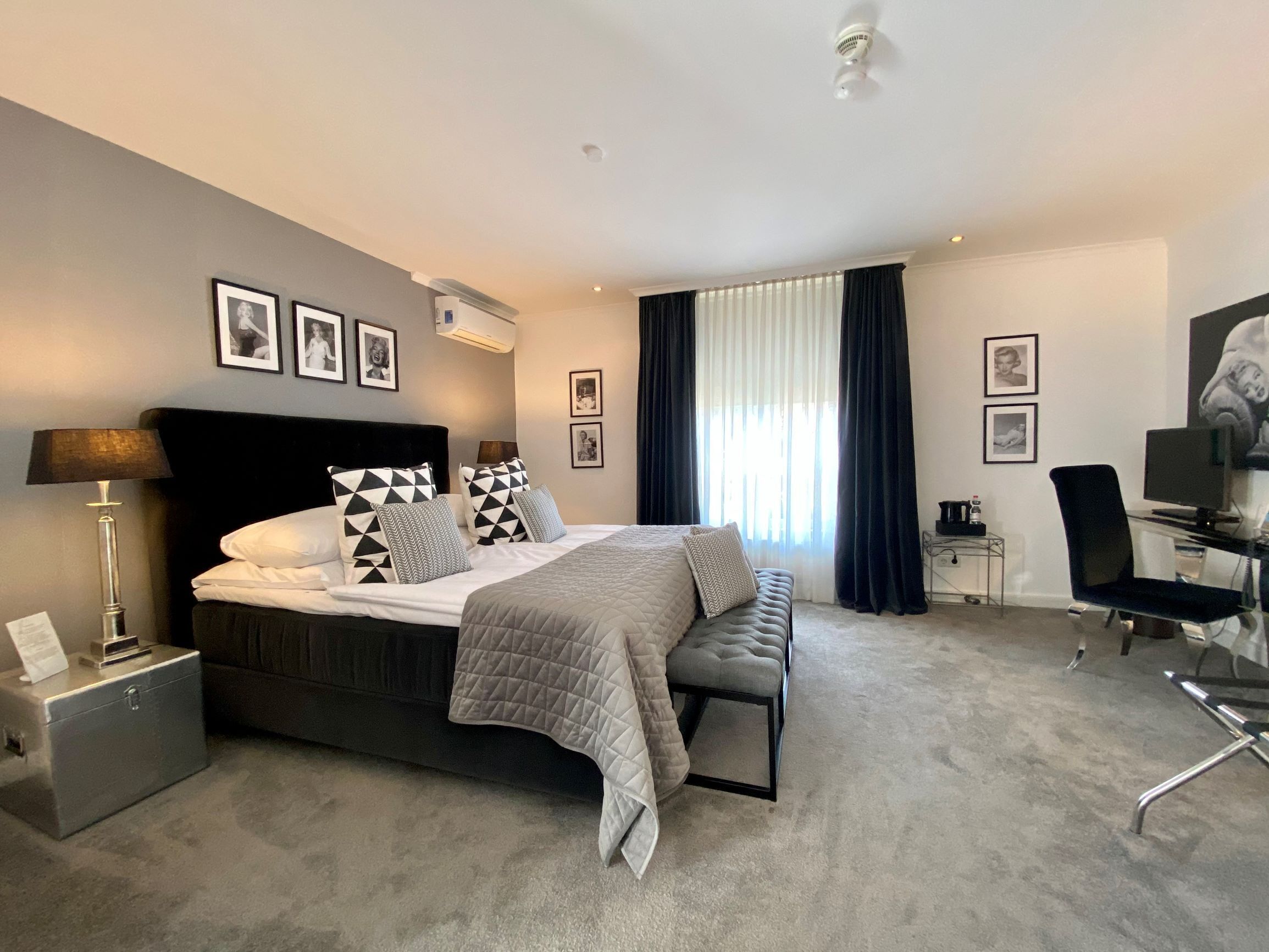Una habitación de hotel moderna con una cama grande, un escritorio y una silla. Hay cuadros enmarcados en la pared y una ventana con cortinas.