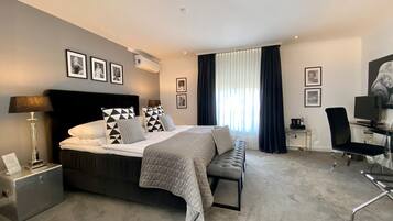 A modern hotel room with a large bed, a desk, and a chair. There are framed pictures on the wall and a window with curtains.