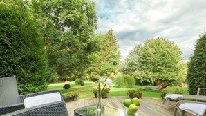 Een terras buiten met rieten meubilair, een glazen tafel en uitzicht op een verzorgde tuin.