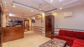 A hotel lobby with a reception desk, a red sofa, a wooden cabinet, a ceiling fan, and a TV mounted on the wall.