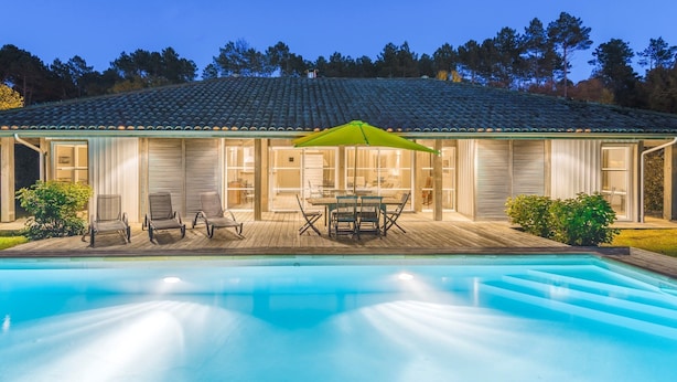 A swimming pool with a wooden deck and lounge chairs, a covered patio with a table and chairs, and a house with a tiled roof and large windows. - Villa avec Piscine Privée | WiFi Gratuit + Parking (Moliets)