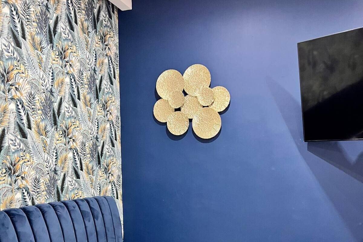 A bedroom with a blue wall, a decorative wall piece, a TV mounted on the wall, a bed with a blue headboard, and a patterned wallpaper.