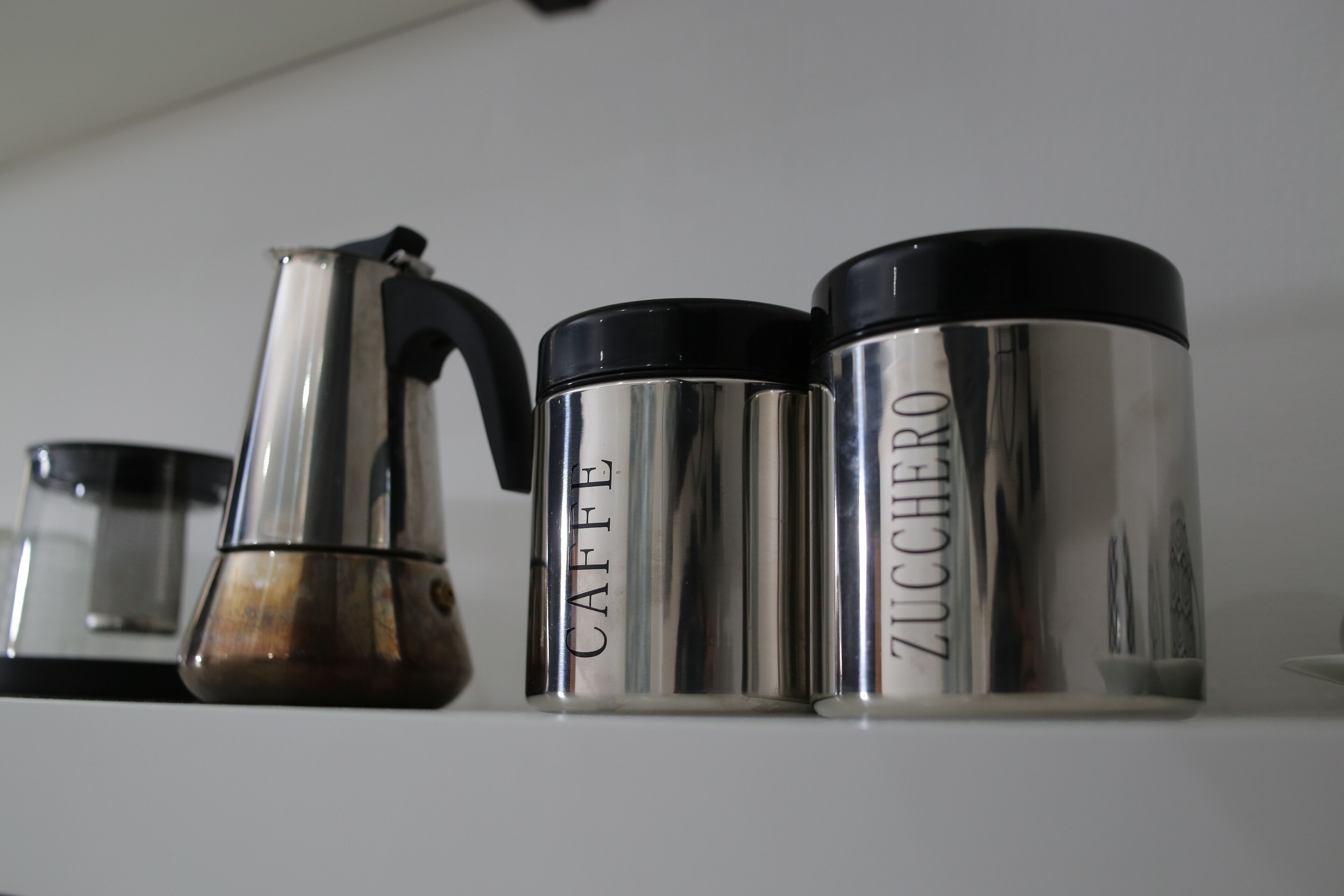 Three stainless steel containers with black lids on a white shelf.