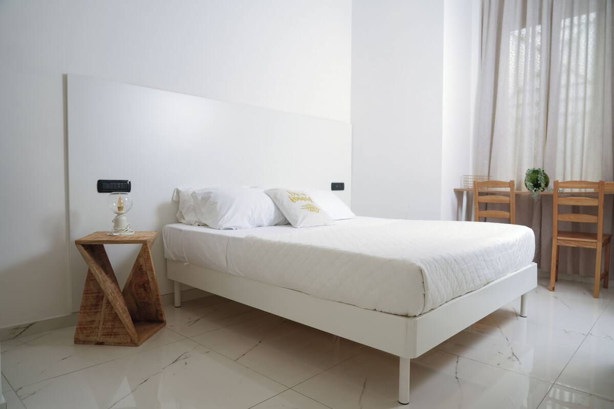 A modern bedroom with a white bed, a wooden bedside table, and a small plant.