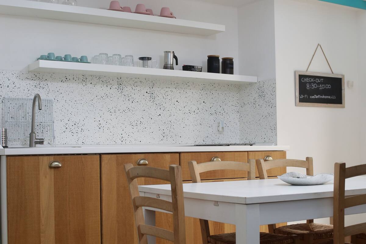 A kitchen with a white countertop, wooden cabinets, and a dining table with chairs. There are shelves above the countertop holding various kitchen items.