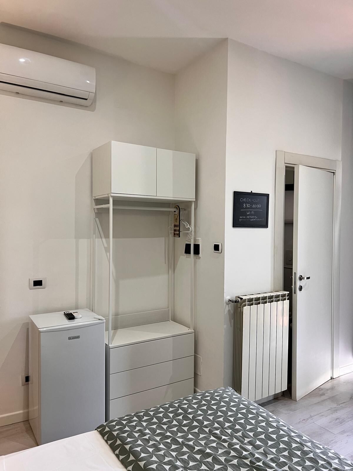Una habitación pequeña con cama, mesita de noche, armario, radiador y una puerta con un cartel.