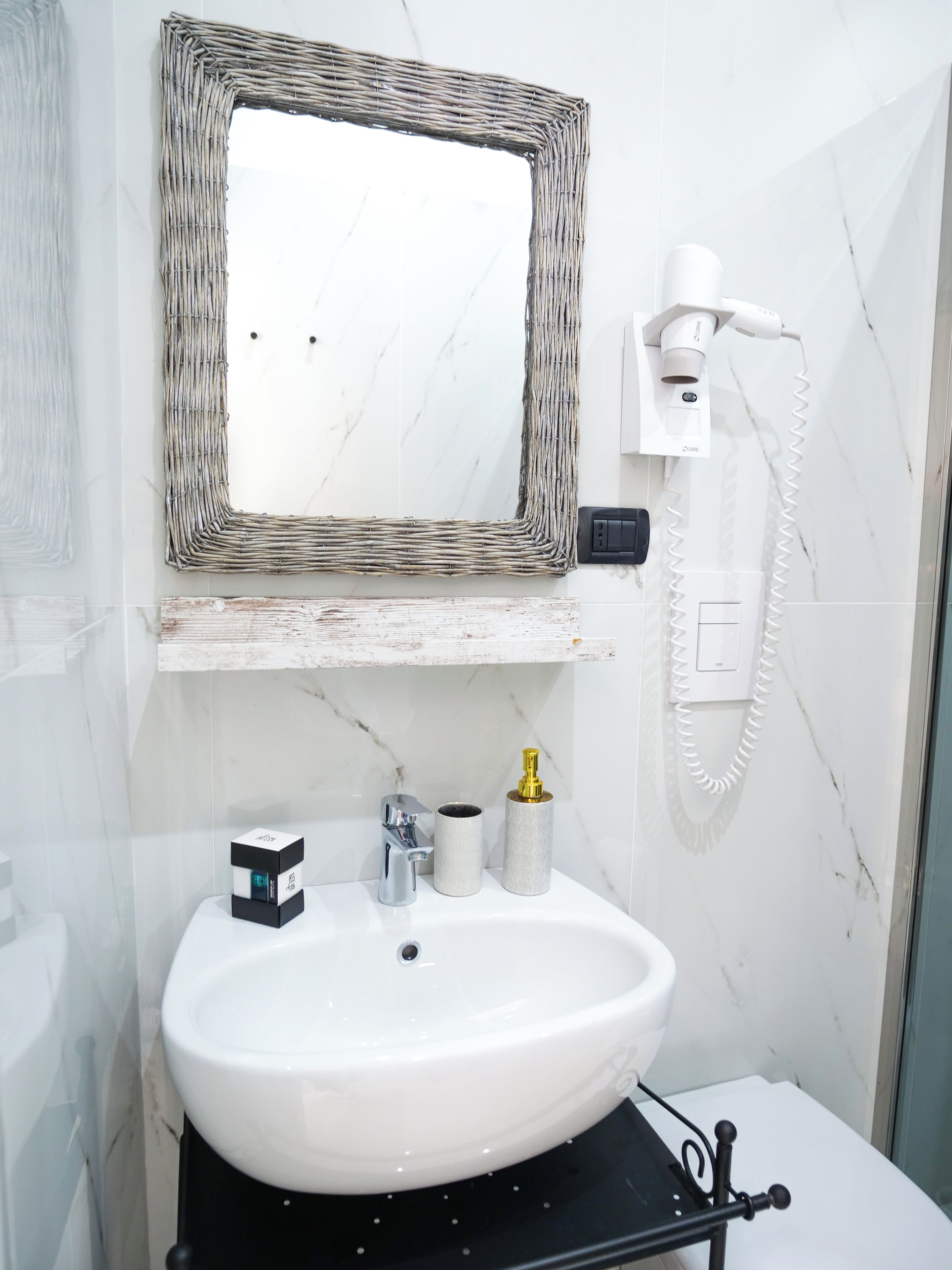 A bathroom with a white marble wall, a white sink, a chrome faucet, a white toilet, a wicker-framed mirror, a hairdryer, and a soap dispenser.