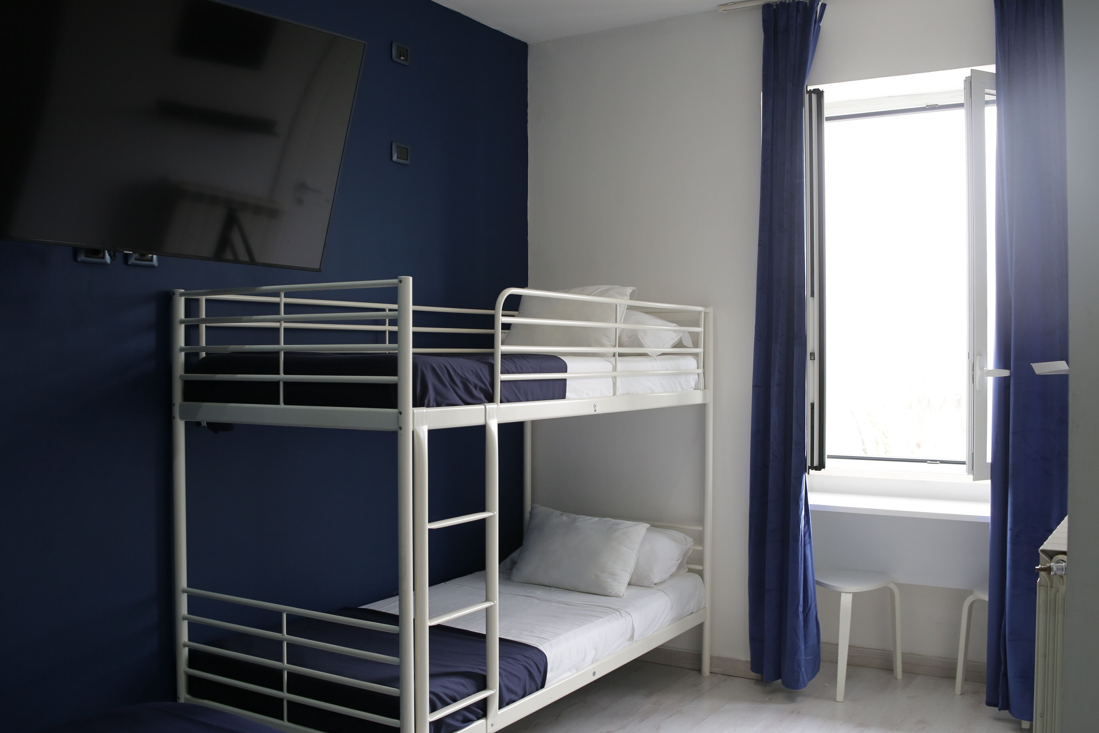 A bunk bed room with a TV on the wall, a window with curtains, and a small white chair.