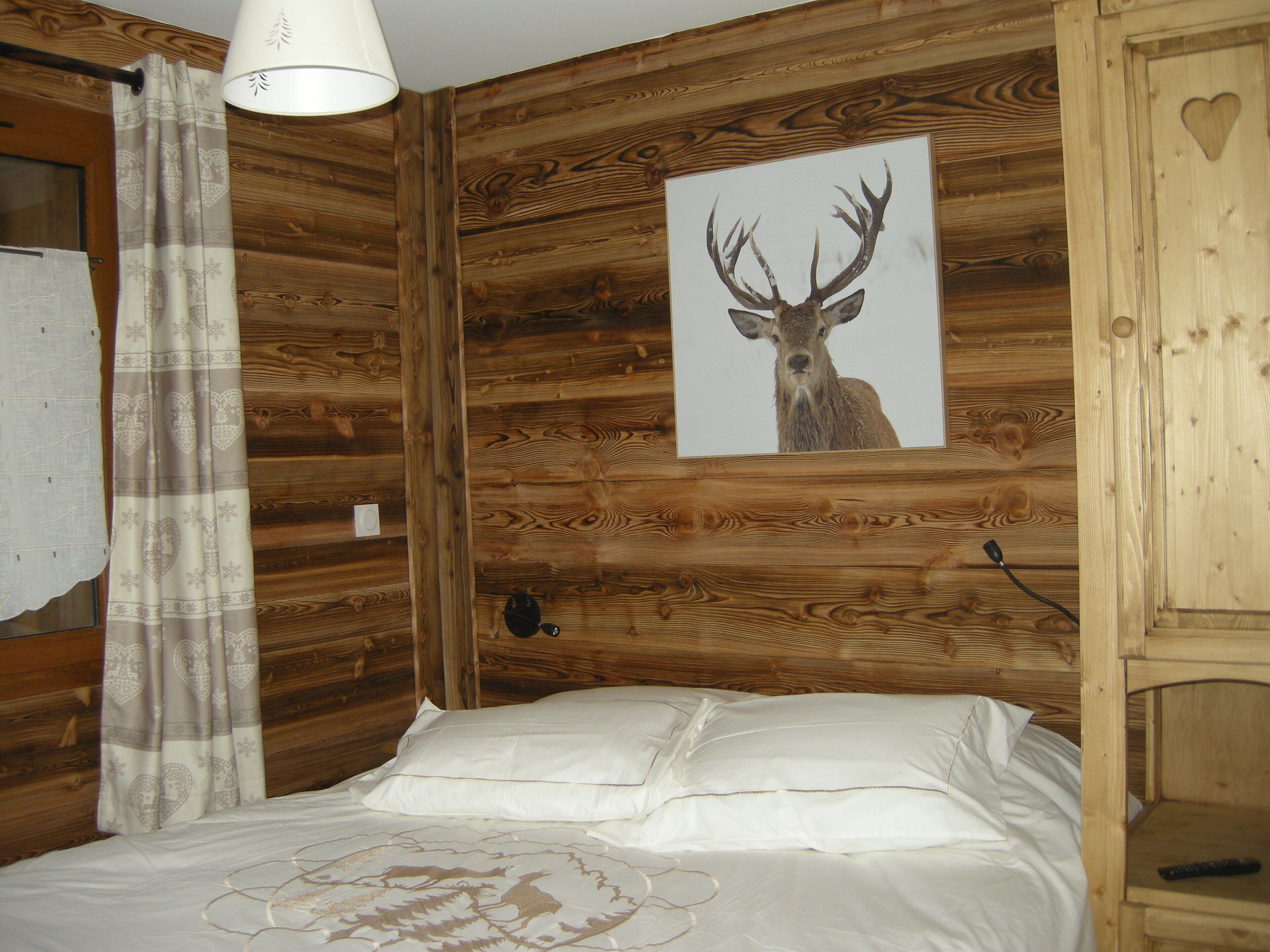 Una habitación tipo cabaña de madera con cama, una lámpara fijada a la pared y una fotografía enmarco de un ciervo.
