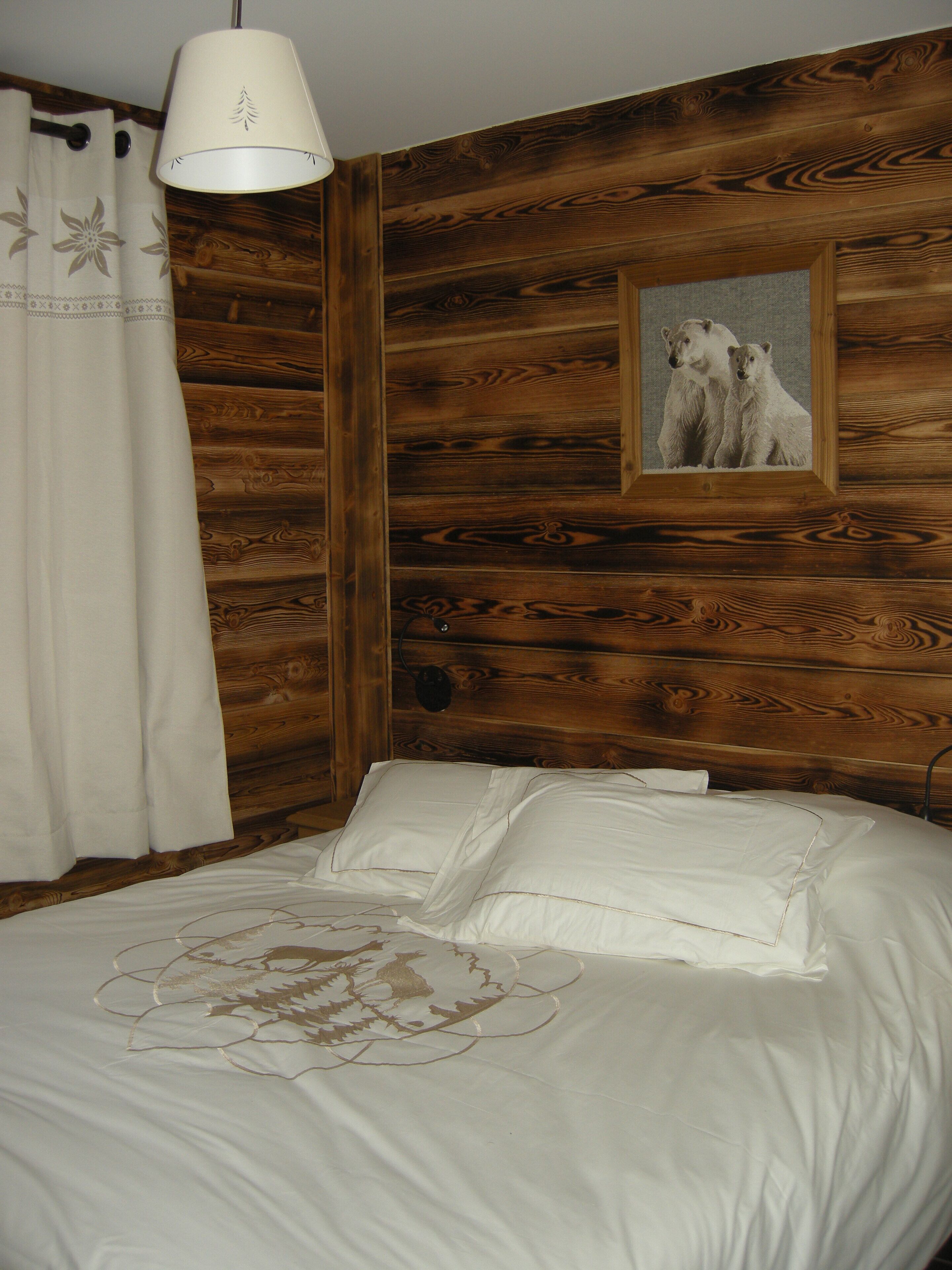 A bedroom with a wooden wall, a bed with white bedding, a white curtain, and a framed picture of bears.