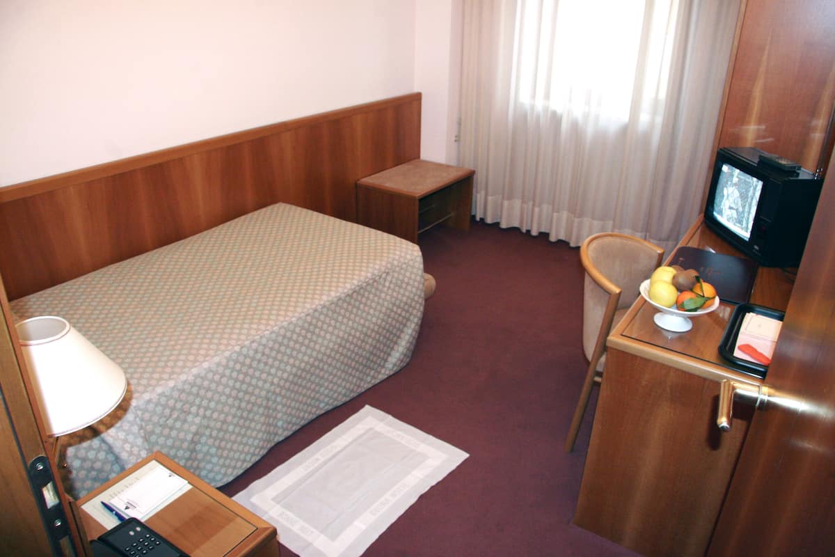 A hotel room with a single bed, a small desk with a television, a chair, and a side table with a lamp and a telephone.