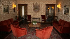 Una habitación con papel tapiz estampado, un reloj grande y ornamentado, y muebles tapizados en rojo.